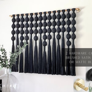 Op de afbeelding: Zwarte macram&eacute; wandhanger met een houten deuvel. Het ontwerp heeft een reeks geknoopte bollen en lange kwasten. De afbeelding toont ook twee hardware-opties: geborsteld satijn messing of matzwart.
