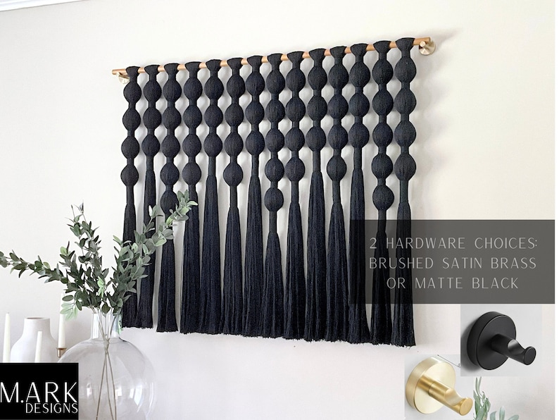 Op de afbeelding: Zwarte macram&eacute; wandhanger met een houten deuvel. Het ontwerp heeft een reeks geknoopte bollen en lange kwasten. De afbeelding toont ook twee hardware-opties: geborsteld satijn messing of matzwart.