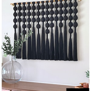 K&ouml;nnte beinhalten: Schwarze Makramee-Wandbehang mit einem Holzstab und schwarzen Quasten. Der Wandbehang zeigt eine Reihe gro&szlig;er, runder Knoten.