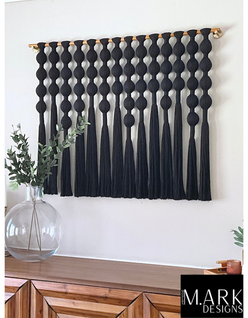 K&ouml;nnte beinhalten: Schwarze Makramee-Wandbehang mit einem Holzstab und schwarzen Quasten. Der Wandbehang zeigt eine Reihe gro&szlig;er, runder Knoten.