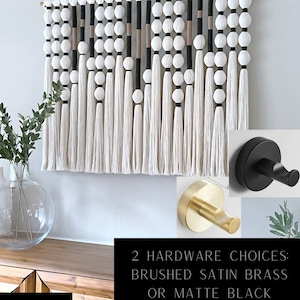 K&ouml;nnte beinhalten: Ein wei&szlig;er, schwarzer und beigefarbener Makramee-Wandbehang mit einem Holzstab und Quasten. Zwei Optionen f&uuml;r Wandhaken werden gezeigt: geb&uuml;rstetes Satin-Messing und mattschwarz. "2 Hardware Choices: Brushed Satin Brass or Matte Black" steht in schwarzer Schrift auf wei&szlig;em Hintergrund.