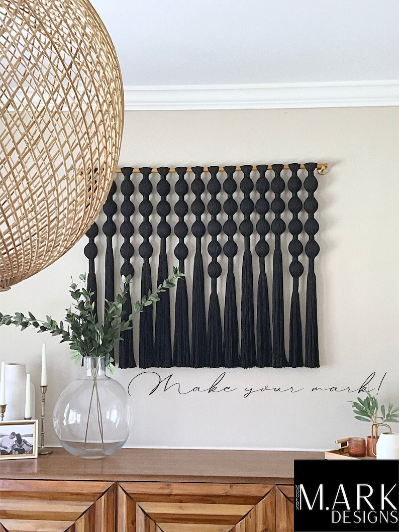 Op de afbeelding: Een zwarte macram&eacute; wandhanger met een houten deuvel en lange kwasten. De wanddecoratie wordt boven een houten kast getoond. De zin "Make your mark!" staat op de muur. Een grote glazen vaas met groen staat op de kast.