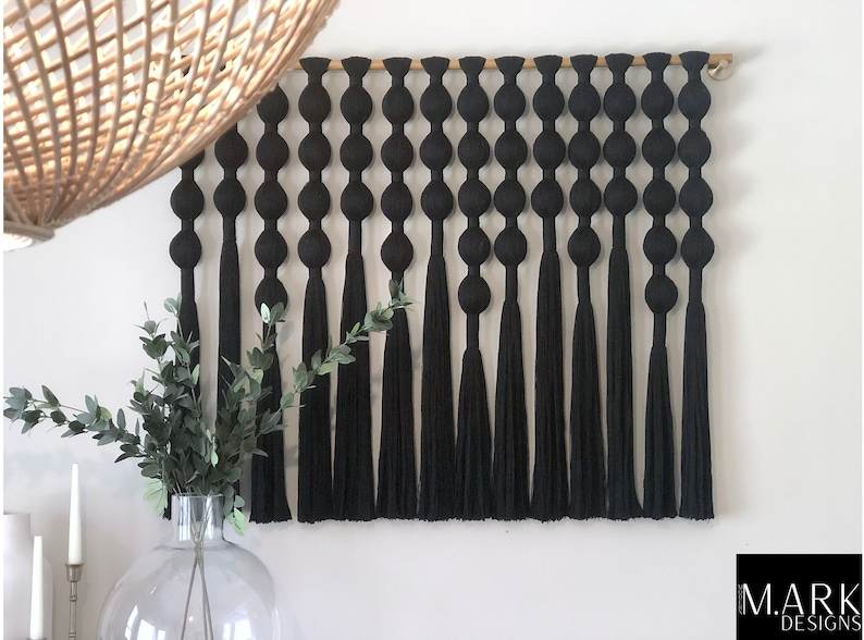 Op de afbeelding: Zwarte macram&eacute; wandhanger met een reeks verbonden, afgeronde vormen en lange kwasten. De wanddecoratie is gemonteerd op een houten roede. Het ontwerp is minimalistisch en modern, geschikt voor diverse interieurstijlen. Het M.A.R.K. DESIGNS-logo staat rechtsonder.