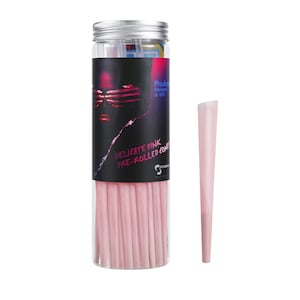 May include: A clear plastic jar filled with delicate pink pre-rolled cones. The jar has a silver lid and a black label with the text "Delicate Pink Pre-Rolled Cones" and a woman's face with her eyes covered by her hands. The label also says "Produced & Designed in NYC". A single delicate pink pre-rolled cone is shown to the right of the jar.