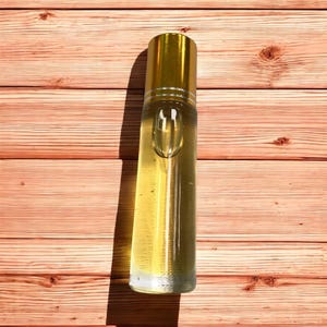 Pode incluir: Uma garrafa de vidro transparente com uma tampa dourada cheia de um l&iacute;quido amarelo claro. A garrafa est&aacute; sobre uma superf&iacute;cie de madeira.