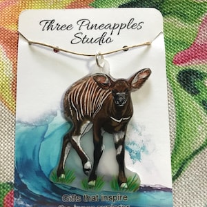 May include: A pendant featuring a detailed illustration of a Kudu, a type of African antelope, with brown and white stripes. The pendant is attached to a white card with the text "Three Pineapples Studio" and "Gifts that inspire the inner explorer."