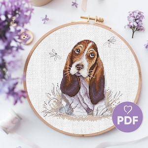 May include: Cross stitch pattern of a brown and white basset hound with long floppy ears, sitting in a field of grass with dragonflies flying around it. The pattern is in a wooden embroidery hoop.