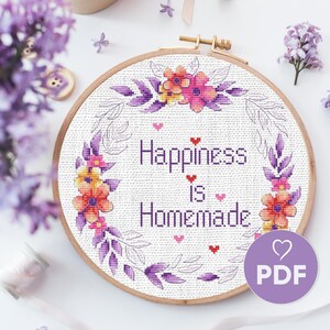 May include: Cross stitch pattern featuring a floral wreath with the text "Happiness is Homemade" in a purple and pink color scheme. The pattern is in a hoop and ready to be stitched.