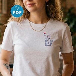 Op de afbeelding: Wit T-shirt met een klein, gepixeld ontwerp van het Vrijheidsbeeld in blauw en rood op de borst. Het T-shirt heeft een ronde hals en korte mouwen. Het ontwerp bevindt zich aan de linkerkant van de borst.