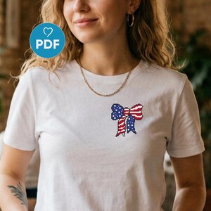 Puede incluir: Camiseta blanca con un diseño de lazo pixelado en rojo, blanco y azul, que recuerda a la bandera estadounidense. La camiseta tiene mangas cortas y cuello redondo. Un icono PDF circular azul es visible en la esquina superior izquierda.