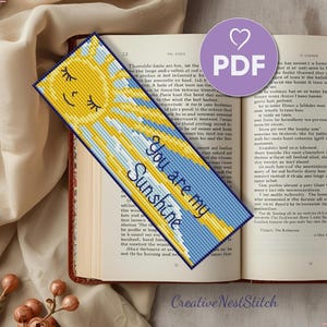 May include: A handmade bookmark with a cross-stitch design. The bookmark features a smiling yellow sun, blue and yellow rays, and the text "You are my Sunshine." It is placed on an open book.