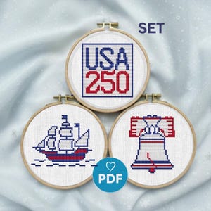 May include: A set of three cross-stitch designs in wooden hoops. The designs include the text "USA 250", a ship, and the Liberty Bell, all in red, white, and blue. The word "SET" is above the hoops.
