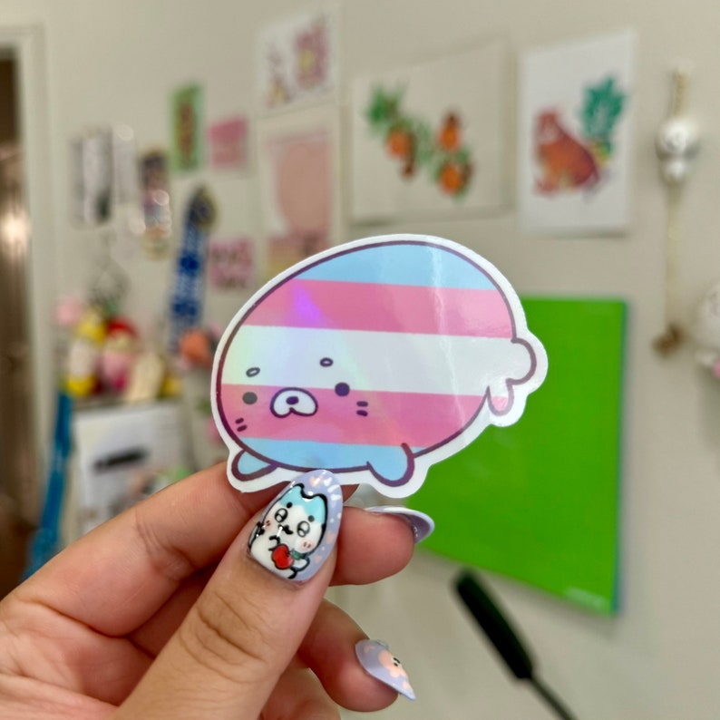 May include: A holographic sticker of a cartoon seal with a transgender pride flag design. The seal is light blue with a white belly and has a pink nose and black eyes.