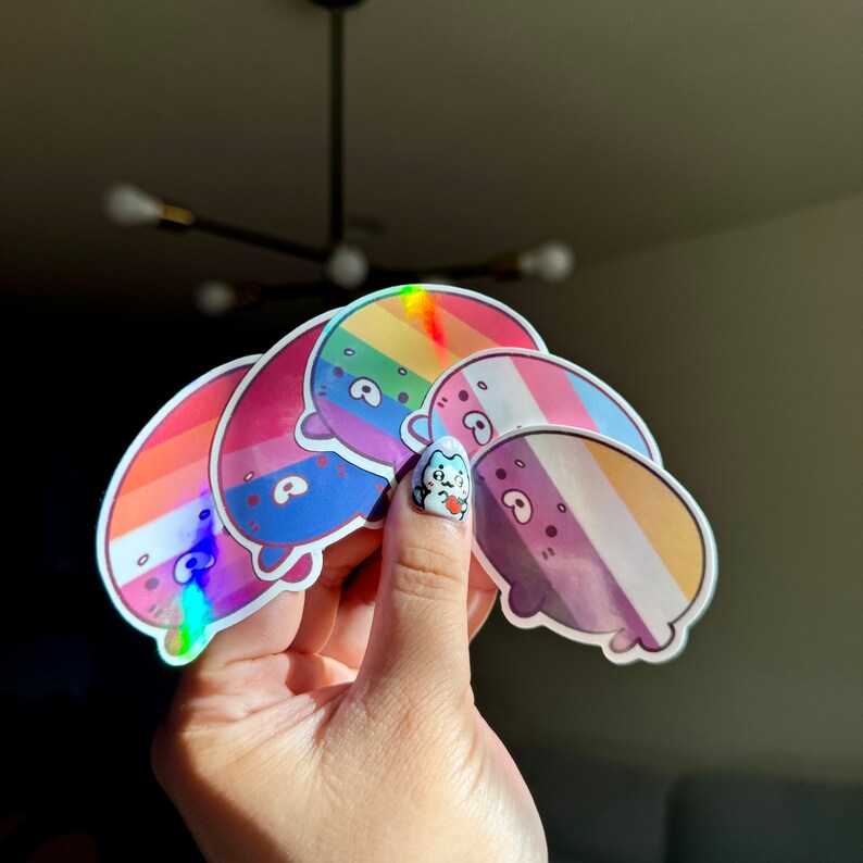 May include: A set of five iridescent stickers featuring cartoon characters with different pride flag designs. The stickers are holographic and have a rainbow, bisexual, transgender, non-binary, and lesbian pride flag design.