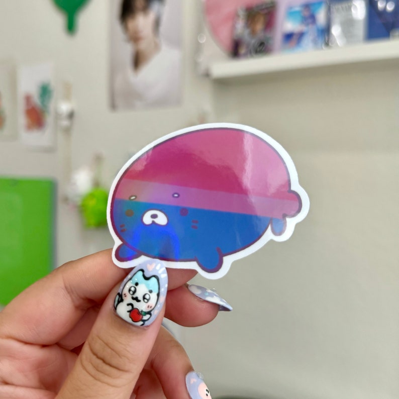 May include: A holographic sticker of a cartoon bear with a pink, blue, and white color scheme. The bear is lying on its side with its head turned to the left. The sticker is on a white background.