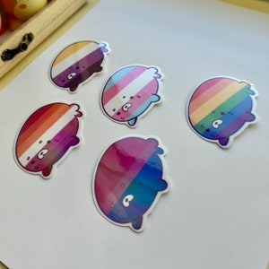 May include: Five round stickers with different pride flag designs. Each sticker features a cartoon bear with a round body and small legs and arms. The stickers are on a white background.