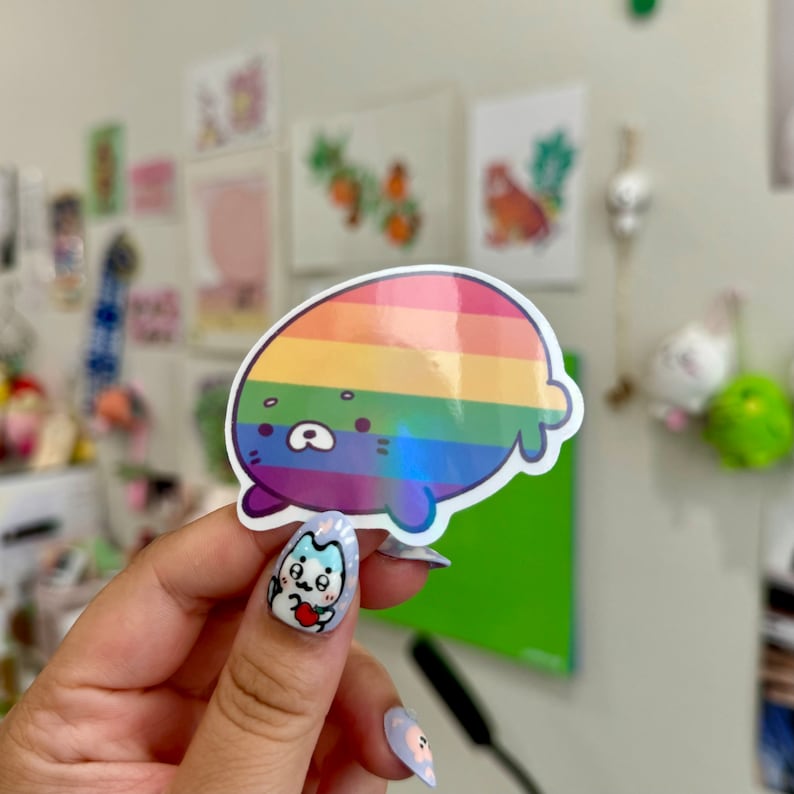 May include: A rainbow-colored sticker of a cartoon seal with a holographic finish. The seal is smiling and has a rainbow pattern on its body.