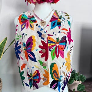 May include: A cream-colored sleeveless vest featuring vibrant, multi-colored butterfly and floral embroidery. The garment has a V-neckline, a front button closure with four light brown buttons, and a textured fabric finish throughout the piece.