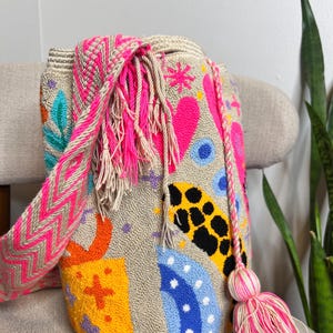 May include: A beige, hand-stitched bag with a colorful design. The bag features a pink and beige chevron strap, a pink tassel, and embroidered shapes in pink, orange, yellow, blue, and black. The bag is sitting on a beige chair.
