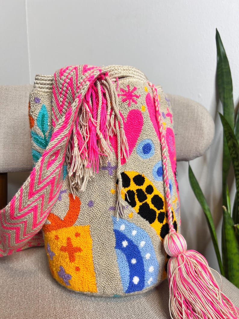 May include: A beige, hand-stitched bag with a colorful design. The bag features a pink and beige chevron strap, a pink tassel, and embroidered shapes in pink, orange, yellow, blue, and black. The bag is sitting on a beige chair.