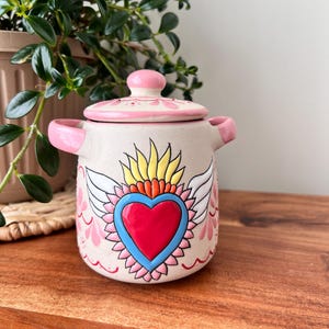 May include: A ceramic pot with a lid, painted with a sacred heart design. The pot is beige with pink handles and a pink lid. The heart is red with a blue outline, surrounded by yellow flames and white wings. Decorative floral accents complete the design.