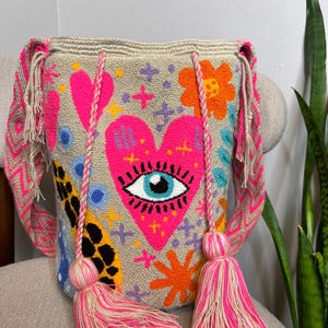 May include: Beige shoulder bag with a vibrant, embroidered design. The bag features a large pink heart with an eye, surrounded by colorful flowers, stars, and other shapes. It has pink and beige straps and tassels, adding a playful touch to the accessory.
