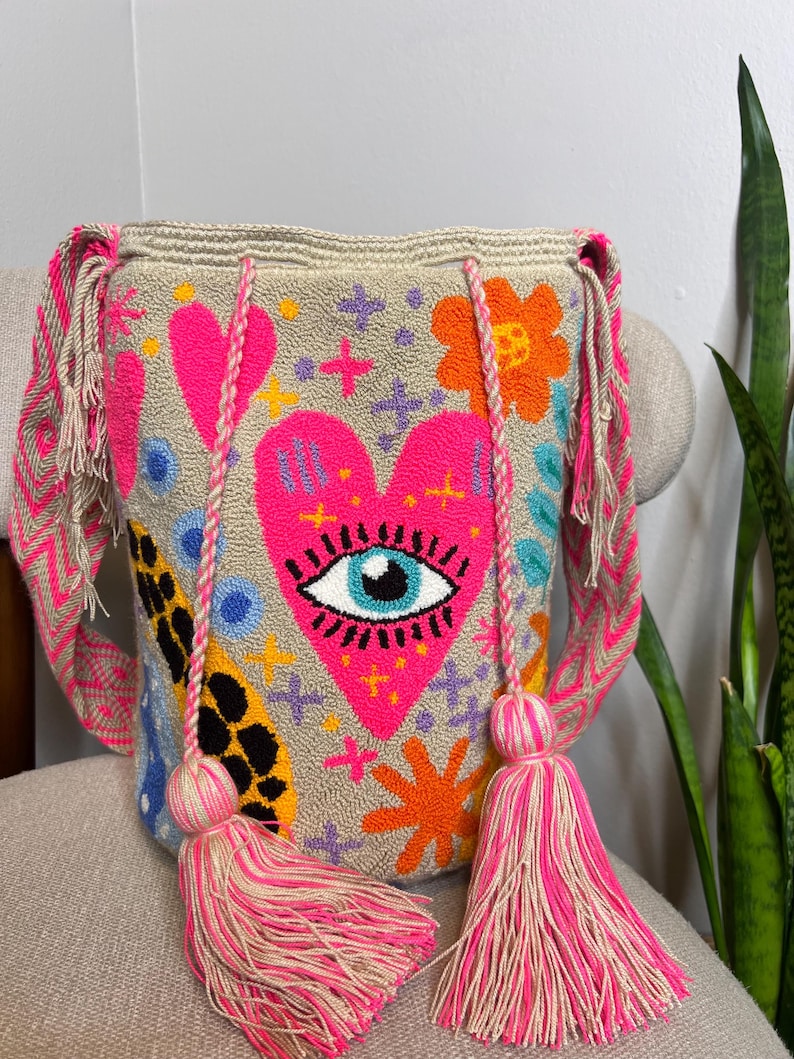 May include: Beige shoulder bag with a vibrant, embroidered design. The bag features a large pink heart with an eye, surrounded by colorful flowers, stars, and other shapes. It has pink and beige straps and tassels, adding a playful touch to the accessory.