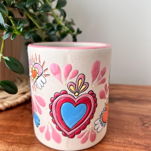 May include: White ceramic mug with a pink rim and a colorful design of hearts, flowers, and wings. The hearts are red, pink, and blue.