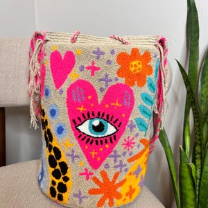 May include: A beige bucket bag with a vibrant, embroidered design. The bag features a large pink heart with an eye, along with other colorful floral and geometric patterns. The bag has pink and beige fringe and rope handles.