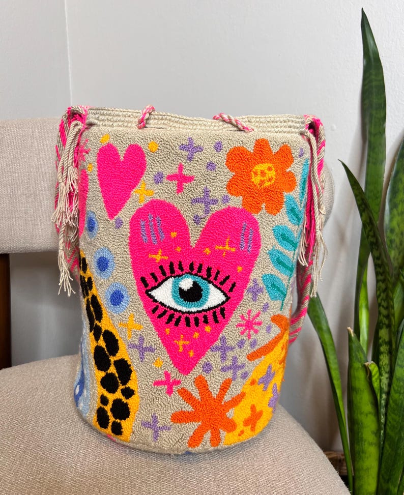 May include: A beige bucket bag with a vibrant, embroidered design. The bag features a large pink heart with an eye, along with other colorful floral and geometric patterns. The bag has pink and beige fringe and rope handles.