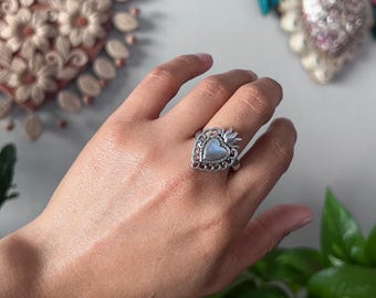 May include: A silver-toned ring featuring a central heart-shaped iridescent stone set within an ornate, filigree-style frame that includes a small flame-like detail at the top, showcasing intricate metalwork and a polished finish.