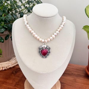 May include: A pearl necklace with a heart-shaped pendant. The pendant has a red stone center and a silver filigree design. The necklace is displayed on a white bust against a background of greenery.