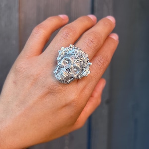 May include: A silver-toned ring featuring a detailed skull design adorned with sculpted roses and a small cross symbol on the forehead. The metal surface has an oxidized finish to highlight the intricate textures of the floral and bone elements.