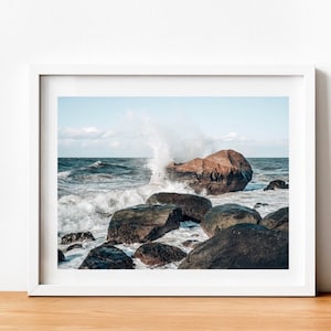 May include: A large, brown rock sits in the ocean with waves crashing against it. The water is a deep blue and the sky is a light blue with white clouds. There are smaller rocks in the foreground.