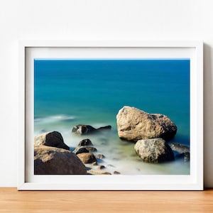 May include: A photograph of a rocky shoreline with large, smooth rocks in the foreground. The rocks are partially submerged in the water, which is a light blue color and appears to be moving. The background is a hazy blue sky.