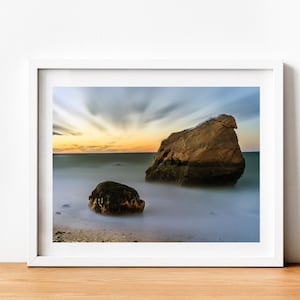 May include: A photograph of two large rocks on a beach at sunset. The rocks are brown and gray, and the water is a light blue. The sky is a soft orange and pink.