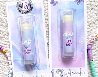 Puede incluir: Dos brillos labiales en envases transparentes con el texto "Ariah's" y "13" en la parte delantera. Uno dice "Birthday Version Balm" y el otro "Best Era Balm". Pulseras de cuentas de colores rodean los brillos labiales.