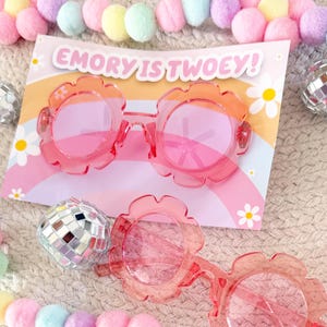 May include: Pink flower-shaped sunglasses with a party invitation that reads "EMORY IS TWOEY!". The sunglasses are set on a light-coloured surface with decorative pom-poms and disco balls.