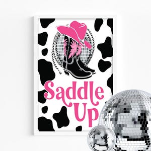 Puede incluir: Impresión enmarcada con un sombrero de vaquero rosa y botas de vaquero negras, una bola de discoteca y las palabras "Saddle Up" en rosa. El fondo tiene un estampado de vaca en blanco y negro.