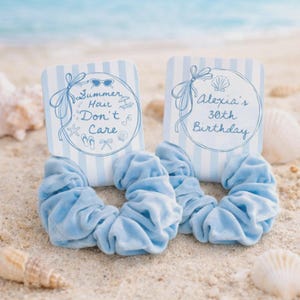 Op de afbeelding: Twee lichtblauwe scrunchies met bijpassende kaarten op zand. De kaarten hebben blauw-witte strepen en bevatten tekst en illustraties. E&eacute;n kaart zegt "Summer Hair Don't Care". De andere zegt "Alexia's 30th Birthday".