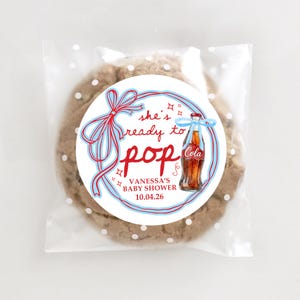 Pu&ograve; includere: Un biscotto con gocce di cioccolato in un sacchetto di cellophane trasparente con pois bianchi. Il biscotto ha un adesivo circolare con il testo "she's ready to pop" e un'illustrazione di una bottiglia di cola vintage. L'adesivo include anche il testo "VANESSA'S BABY SHOWER 10.04.26".