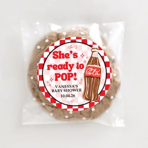 Peut inclure: Un cookie aux p&eacute;pites de chocolat dans un sac en cellophane transparent &agrave; pois blancs. Le cookie porte un autocollant circulaire avec le texte "She's ready to POP!" et un dessin de bouteille de Coca-Cola. L'autocollant comprend &eacute;galement le texte "VANESSA'S BABY SHOWER 10.04.26".