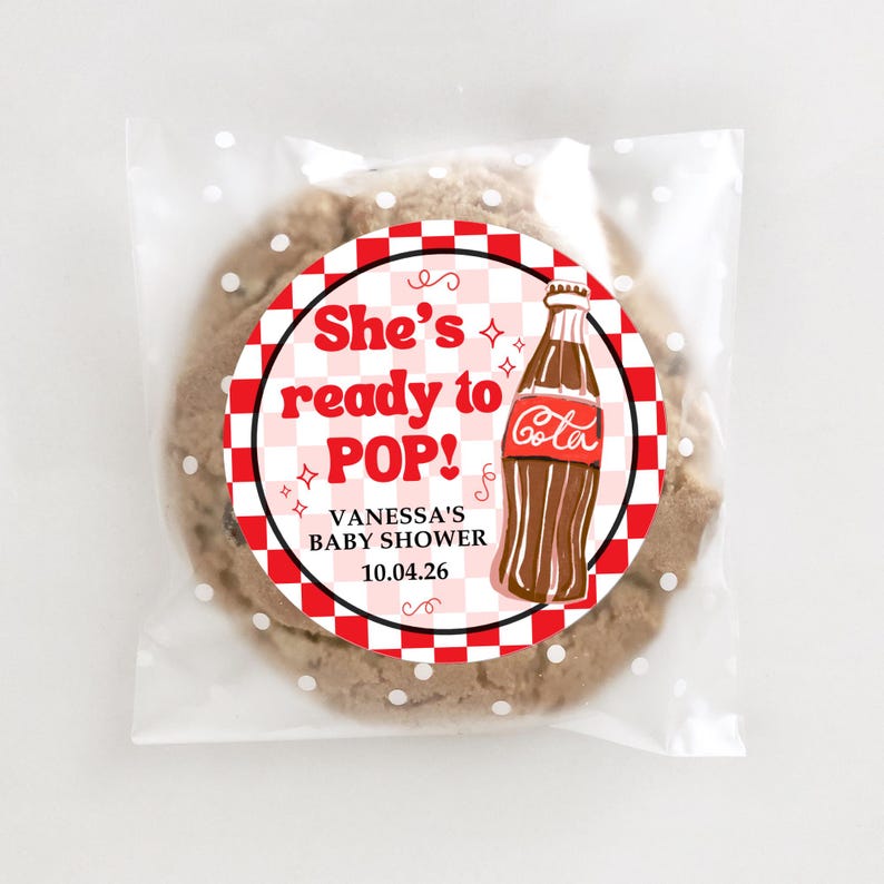 Peut inclure: Un cookie aux p&eacute;pites de chocolat dans un sac en cellophane transparent &agrave; pois blancs. Le cookie porte un autocollant circulaire avec le texte "She's ready to POP!" et un dessin de bouteille de Coca-Cola. L'autocollant comprend &eacute;galement le texte "VANESSA'S BABY SHOWER 10.04.26".