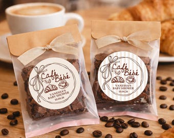 May include: Two clear cellophane bags filled with brownies, each sealed with a brown paper flap and tied with a beige ribbon. Each bag has a round sticker with the text "Café Bébé" and "Vanessa's Baby Shower 10.04.26". Coffee beans and a cup of coffee are in the background.