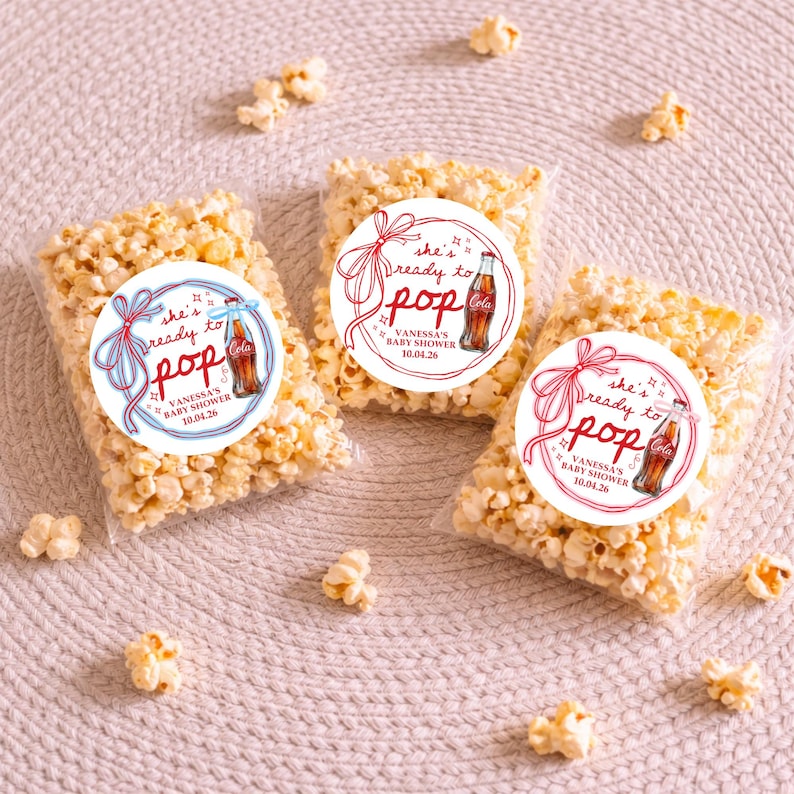 Pu&ograve; includere: Tre sacchetti trasparenti pieni di popcorn, ciascuno adornato con un adesivo circolare. Gli adesivi presentano il testo "she's ready to pop" con un'illustrazione di una bottiglia di Coca-Cola e il testo "VANESSA'S BABY SHOWER 10.04.26".