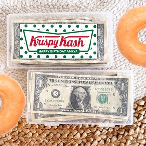 May include: Two clear plastic containers filled with dollar bills. One container has a label that reads "Krispy Kash" and "Happy Birthday Amaya." Two orange donuts are on the sides.