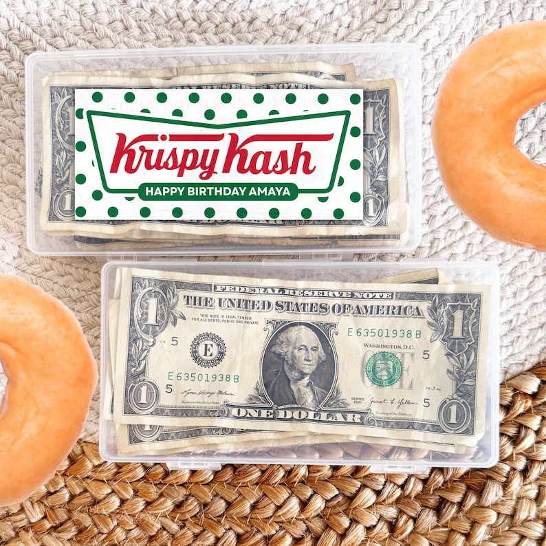 May include: Two clear plastic containers filled with dollar bills. One container has a label that reads "Krispy Kash" and "Happy Birthday Amaya." Two orange donuts are on the sides.