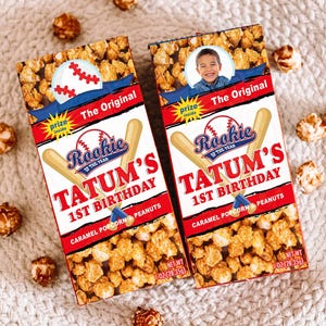 May include: Two boxes of caramel popcorn with baseball-themed graphics. The boxes read "Rookie of the Year" and "Tatum's 1st Birthday." Each box has a baseball and a photo of a smiling child. The boxes are filled with popcorn and peanuts.