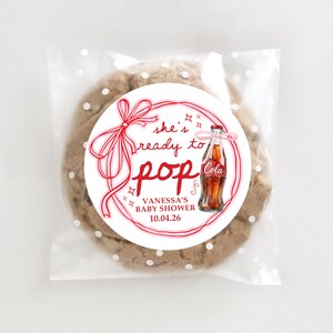Pu&ograve; includere: Un biscotto con gocce di cioccolato in un sacchetto di cellophane trasparente con pois bianchi. Un adesivo rotondo con testo rosso e un'illustrazione di bottiglia di cola recita "she's ready to pop" per il baby shower di Vanessa il 10.04.26.