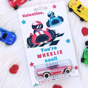 May include: A Valentine's Day card with a red and white toy car in a clear plastic case. The card features race cars and the text "You're WHEELIE cool! cool!" surrounded by stars. Several toy cars and red heart-shaped candies are scattered around the card.